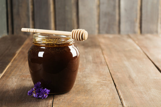 A glass jar of Granny's Krud Kutter Syrup with a wooden honey dipper on top, placed on a rustic wooden table, with a purple flower beside it and a wooden backdrop. This syrup is sourced from Crushed Botanicals Apothecary.