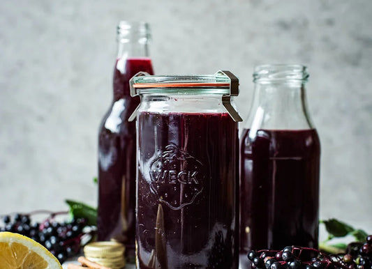 Three containers of Crushed Botanicals Apothecary Black Elderberry Syrup with a rustic background include a glass jar with a clamp lid, a bottle, and a jug accompanied by fresh lemons and blackberries.