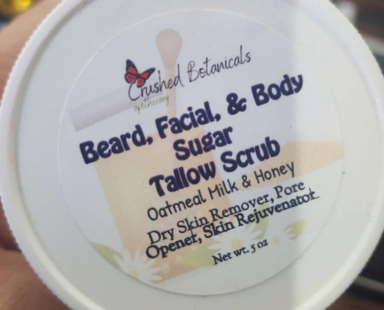 A close-up image of a container lid for a Crushed Botanicals Apothecary product. The label reads "Grass-Fed Tallow Beard, Facial, and Body Scrub, Oatmeal Milk & Honey with North Georgia Grass-Fed Tallow." It claims benefits like "Dry Skin Remover, Pore Opener, Skin Rejuvenator." Net weight is 5 oz.